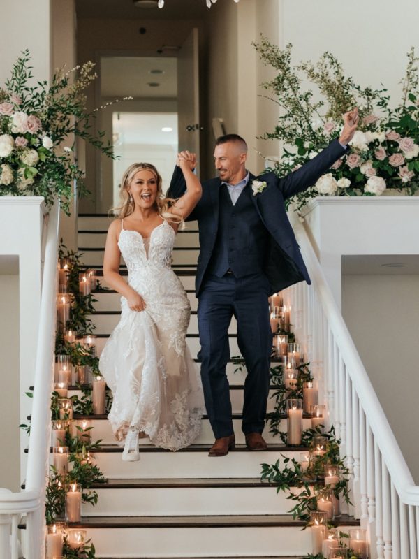 bridge and groom walking downstairs of the wedding venue with wedding floral arrangements lining the stairs